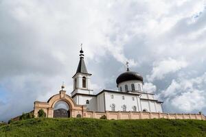 el blanco iglesia, el monasterio puertas, el ladrillo muro, el campana torre, el ortodoxo Iglesia soportes en el montaña en contra el antecedentes de el cielo. foto