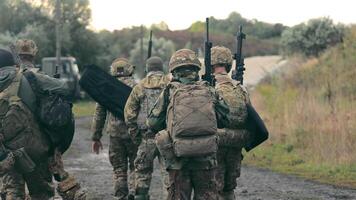 A group of military men returning after completing a task. Soldiers in military uniform with machine guns walk on a stony road with their backs to the cell. video