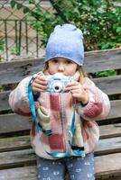 A young girl is sitting on a rustic wooden bench in a park, taking pictures with a blue camera. She is wearing a warm winter coat and a blue beanie hat. The background is a green leafy blur. photo