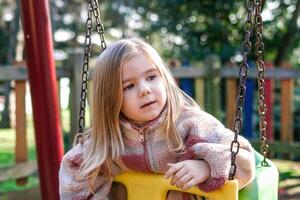 A young girl sits on a swing in a retro-styled playground, her expression is thoughtful and introspective. Her blonde hair falls in waves around her face, and she wears a cozy, casual outfit. photo