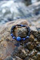 Bangle made from natural materials - black agate, blue lazurite and wooden elements on a wet stone near to sea. Handcraft precious item. Jewelry accessories. photo