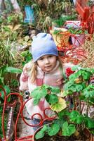 Blonde girl in a cozy jacket and blue hat standing next to a flower pot filled with red flowers. The background features lush greenery and red chairs, creating a beautiful and vibrant scene outdoors. photo