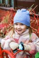 A young girl with blonde hair smiles in a blue hat with white polka dots, a colorful jacket, and white and pink gloves. Isolated on a red background, the image exudes joy. photo