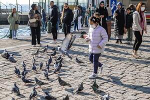 Istanbul, Turkey - December 29, 2022. Adorable Little Girl in White Jacket Walking Among Pigeons in Vibrant City Square on a Sunny Day photo