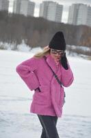 de moda caucásico niña en un caliente rosado invierno falso piel abrigo, negro sombrero y Gafas de sol, hablando en un móvil teléfono en contra el fondo de nieve en un parque en invierno. foto