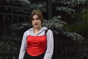 A slender young Caucasian girl in a stylish white shirt and red top against a background of trees. photo