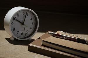 Selective focus at white table clock and ballpoint pen on stack notebooks with sunlight and shadow on wooden table surface photo