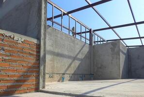 Concrete wall structure inside of room with metal shed roof framing under construction against blue sky background in low angle and perspective side view photo