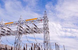 High voltage electric pylons with insulators and equipment of power distribution building station against clouds on blue sky, low angle view with copy space photo