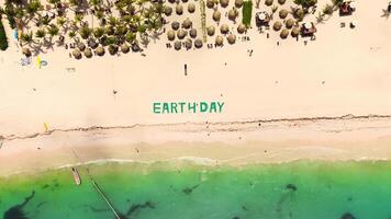 Aerial top view of spacious green sign on tropical beach displaying EARTH DAY, individuals strolling along sandy shore. Setting includes Caribbean Sea, beach umbrellas, loungers and luxurious resort video