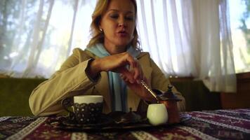 Woman pouring Turkish coffee from cezve into cup. Closeup slow motion shot of female hand with cup on square plate, on table in cafe outdoor. Traditional hot unfiltered coffee served in restaurant video