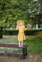 un pequeño niña en un amarillo vestir y Paja sombrero vistiendo un ramo de flores de lilas. un caminar en un primavera parque, cierne lilas. foto