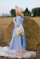 A red-haired woman in a hat and a blue dress walks in a field with haystacks. photo