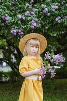 un pequeño niña en un amarillo vestir y Paja sombrero vistiendo un ramo de flores de lilas. un caminar en un primavera parque, cierne lilas. foto