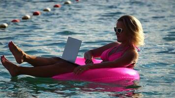 donna libero professionista lavori su il computer portatile nuoto nel mare su rosa gonfiabile squillo. bella signora digitando su computer mentre galleggiante nel il mare su gonfiabile ciambella a tramonto. libero professionista, a distanza opera su vacanza video