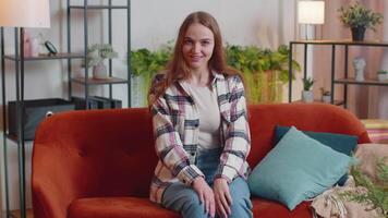 Woman sitting on home sofa, looking at camera, making conference call with friends or parents video