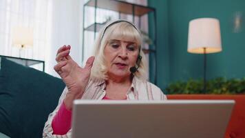 Senior old woman freelance wear headphone make call talking, teaching at laptop at home office video