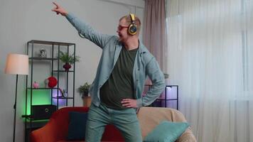 Overjoyed young man in wireless headphones dancing, singing on cozy couch in living room at home video