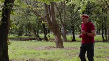 Male senior person running along the road in park. Mature runner man training, listening music video