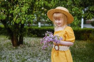 un pequeño niña en un amarillo vestir y Paja sombrero vistiendo un ramo de flores de lilas. un caminar en un primavera parque, cierne lilas. foto