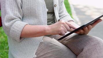 Elderly woman with a tablet in her hands sits on a bench, surfing the internet and researching news on social networks video