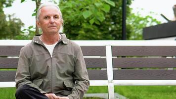 Stylish elderly man with long hair smiling on bench in public city park. Happy senior man looking at camera video