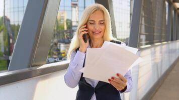 retrato do 50. anos o negócio mulher com documentos chamando telefone ar livre. empresária falando Móvel telefone perto moderno escritório construção video