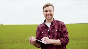 Portrait shot of attractive farmer standing in green field with green wheat and notebook in the hands. Farmer with smile outdoors in summer video