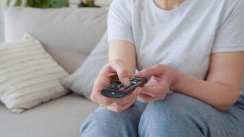 Senior lady watching tv in living room sitting on sofa holding remote control changing tv channels. Senior woman using TV remote control video