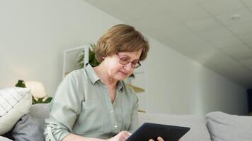 Smiling elderly woman resting on sofa, using digital tablet at home video