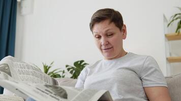 een detailopname visie van een mooi volwassen vrouw is lezing een krant- zittend Aan sofa Bij huis in de ochtend. portret van senior vrouw lezing krant- video