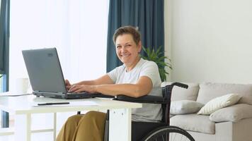 motivata anziano libero professionista è Lavorando digitando nel il computer portatile seduta nel sedia a rotelle a casa. lontano opera concetto video