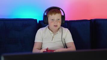 Gamer discussing tactics with teammates while talking into headset. Excited little boy gamer is sitting on a couch, playing and winning in games on console video