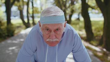 Sénior athlétique homme grand-père fonctionnement le jogging dans ensoleillé parc pendant Matin faire des exercices cardio en plein air video