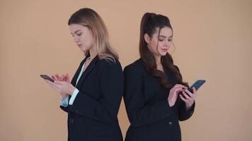 Two young business woman with smartphone standing back to back dressed black suit browsing internet. Technology concept. Always online. One woman shows to friend screen of her smartphone and smiles video