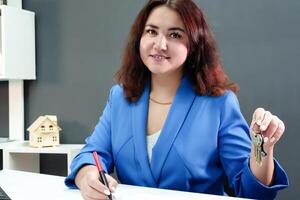 A charming brunette with curly hair and a smile, dressed in a professional blue suit, holds her keys in her hands at her desk, demonstrating her role as a realtor photo