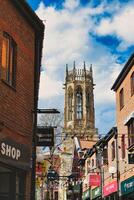 pintoresco urbano calle con festivo verderón líder a un histórico Iglesia torre debajo un azul cielo con mullido nubes en york, norte yorkshire, Inglaterra. foto