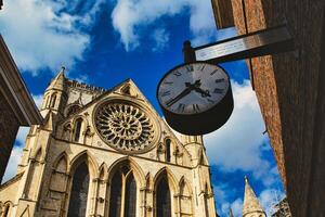 Vintage street clock hanging with a gothic cathedral facade in the background, showcasing intricate architecture and a clear blue sky in York, North Yorkshire, England. photo