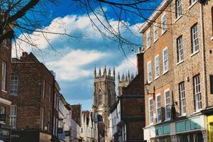 histórico europeo ciudad calle con tradicional ladrillo edificios y un prominente gótico catedral en el antecedentes debajo un azul cielo con nubes en york, norte yorkshire, Inglaterra. foto