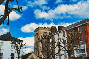 Quaint urban scene with historic stone tower, traditional buildings, and bare tree branches against a vibrant blue sky with fluffy white clouds in York, North Yorkshire, England. photo