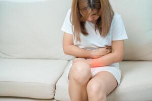 mujer teniendo abdomen dolor debido a estómago dolor, digestión con estreñimiento o Diarrea desde comida envenenamiento, hembra problema y endometriosis, histerectomía, dolor de estómago y menstrual en sofá a hogar foto