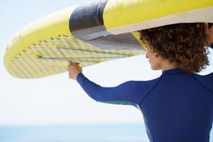 Positive woman in curly hair lifting yellow paddleboard photo