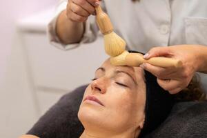 Crop beautician applying cosmetic product on face of female with brushes in beauty salon photo