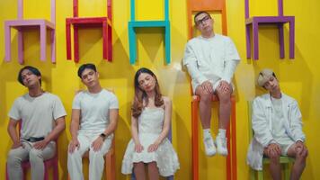 Group of young adults in white attire sitting on colorful chairs against a yellow backdrop, displaying diverse expressions. video