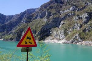 Road sign showing slippery road ahead, next to a cliff with a river below. Careful in driving. Road trip and travel concept. photo