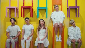 Group of people with diverse expressions sitting and standing against a colorful backdrop with hanging chairs. video