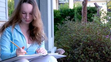 very beautiful teenager girl sitting on porch of house in hands with pencil and white large sketchbook write solve problems online learning do homework sit in park study space for text advertising video