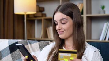 fechar acima sorridente jovem menina segurando Smartphone e bancário crédito cartão, envolvido dentro conectados Móvel compras às lar, feliz fêmea comprador compra bens ou Serviços dentro Internet loja video