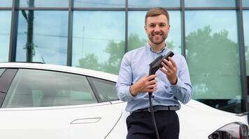 hansome chico en pie cerca su nuevo moderno eléctrico coche y participación enchufe de el cargador video