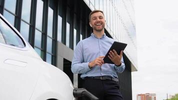 ung europeisk affärsman med trådlös hörlurar och läsplatta nära elektrisk bil, laddning de batteri på stad kraft station video